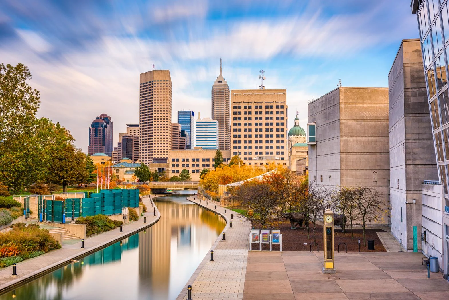 View of a city skyline with a river, autumn trees, and modern buildings under a partly cloudy sky.