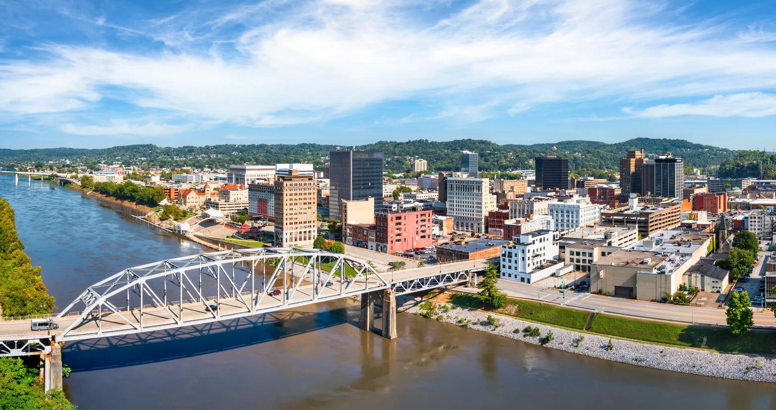 Aerial view of a city skyline with a steel bridge over a river under a partly cloudy blue sky.