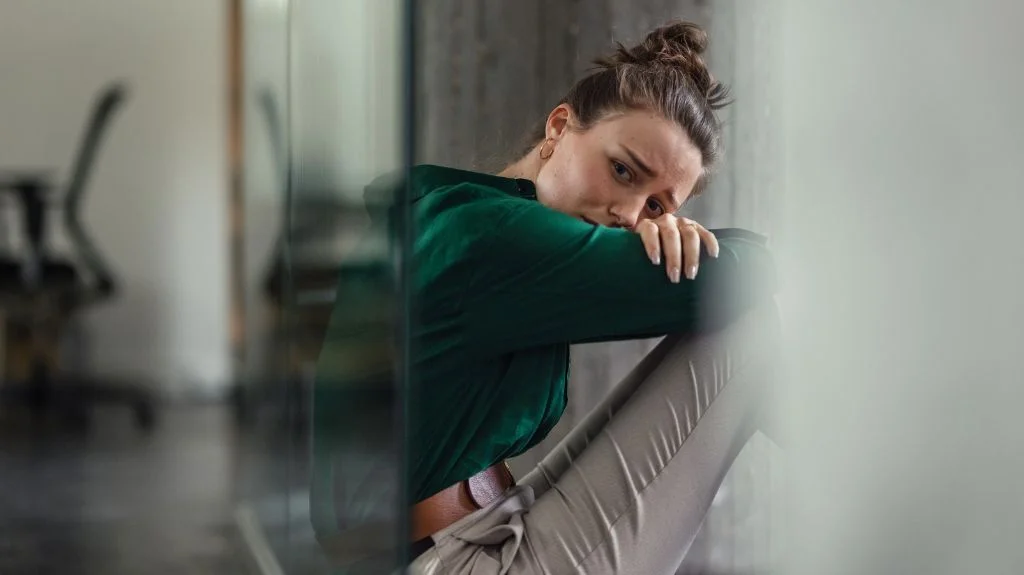 Young woman in green blouse and beige pants sitting on the floor, appearing distressed.