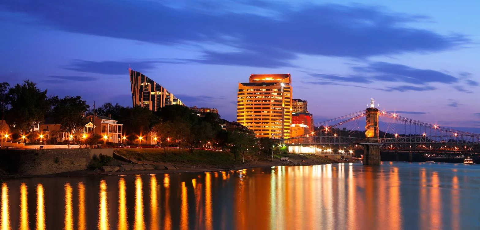 Nighttime cityscape featuring illuminated buildings, a suspension bridge, and water reflections.