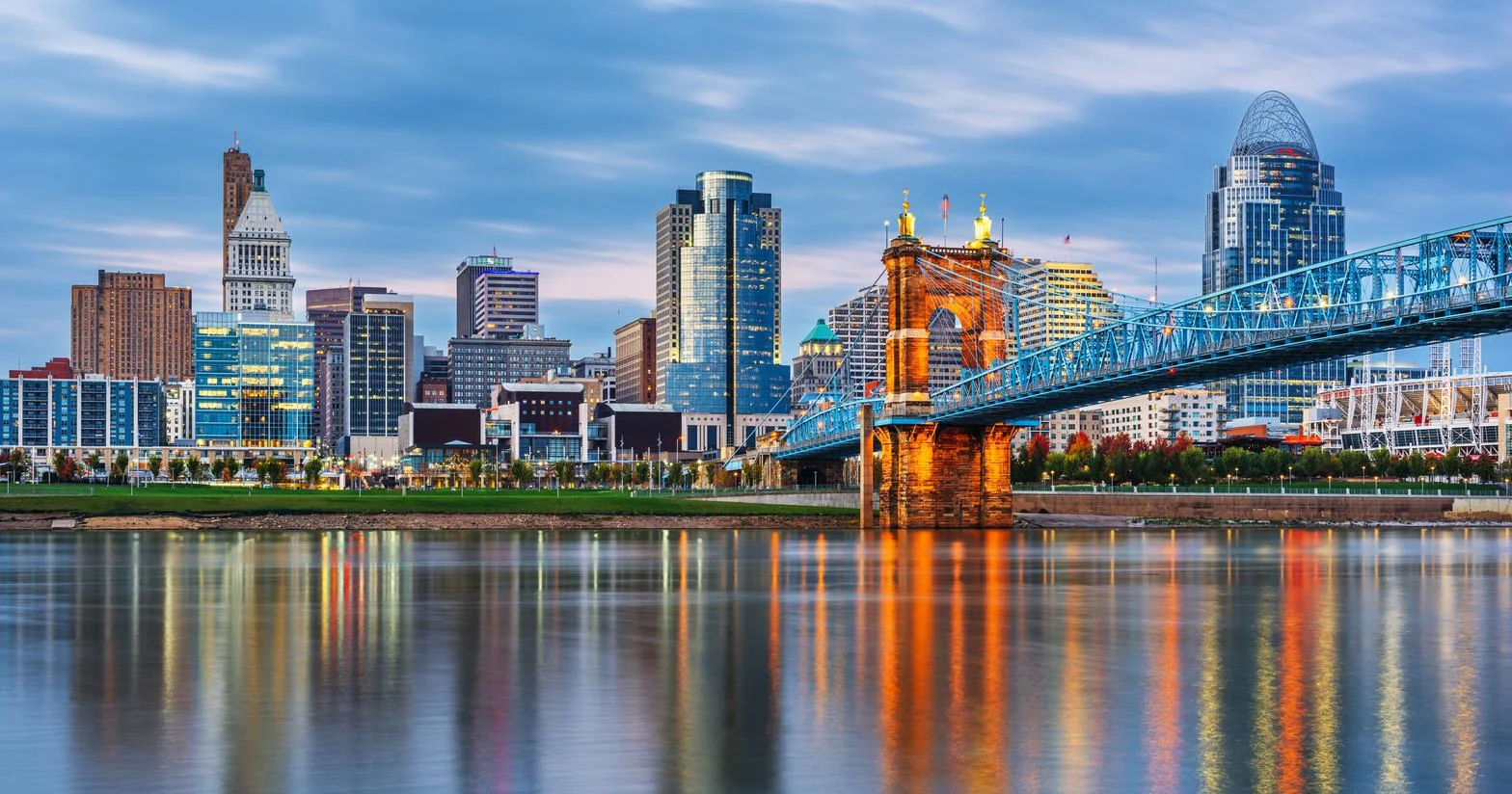 Evening view of Cincinnati skyline with illuminated John A. Roebling Suspension Bridge over calm river.