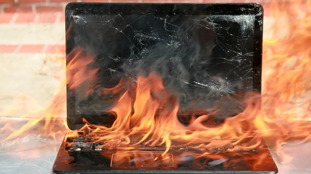 Laptop engulfed in flames with a cracked screen and burning keyboard.