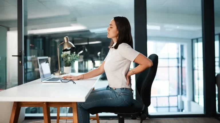 Woman in white shirt and jeans sitting at office desk, holding lower back in discomfort.