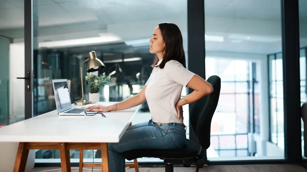 Woman in white shirt and jeans sitting at office desk, holding lower back in discomfort.
