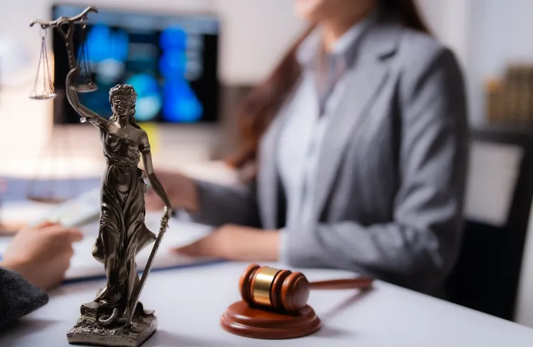 Bronze Lady Justice statue and wooden gavel on desk with blurred woman in gray suit in background.