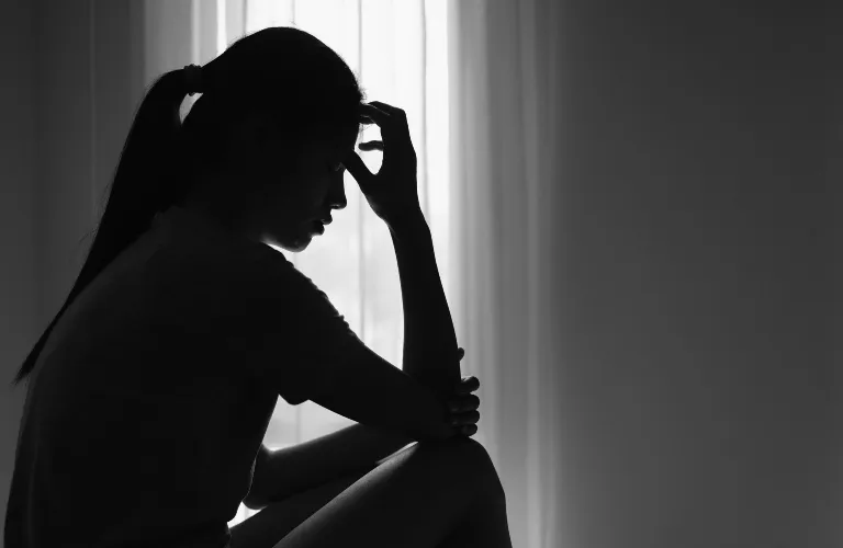 Silhouetted woman sitting indoors near window, holding head in contemplative posture.