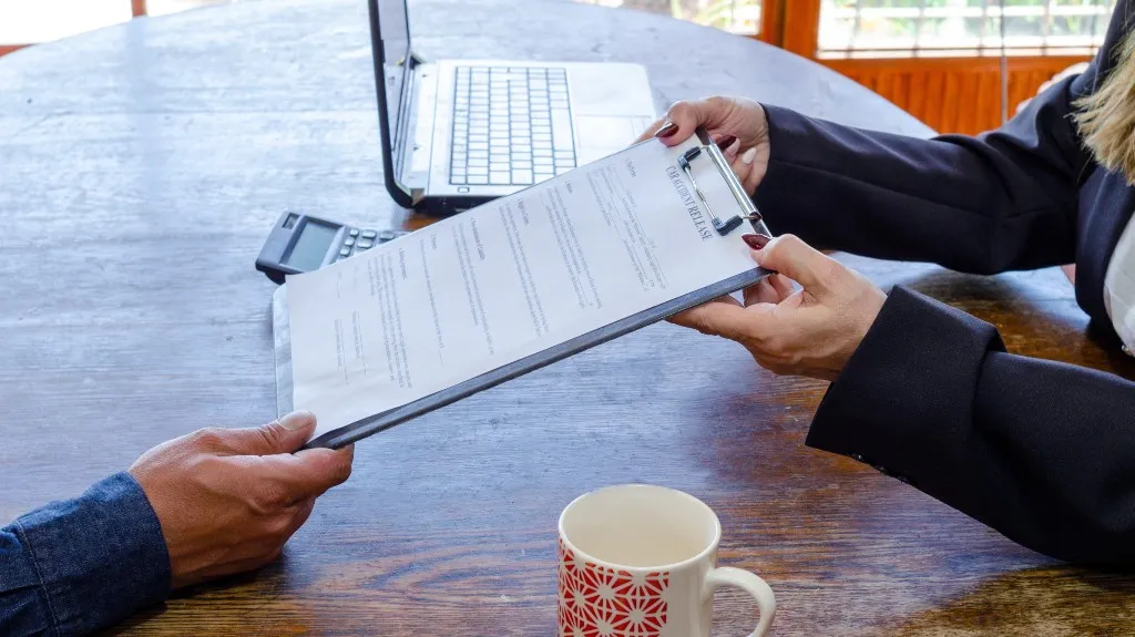 Two people exchanging a clipboard with a contract over a wooden table, laptop and coffee mug visible.