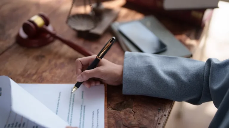 Hand in suit jacket holding pen, signing legal document on wooden desk with gavel and scales.