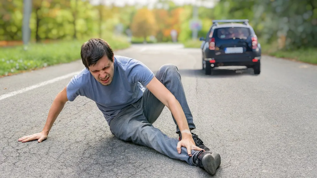 Man in blue shirt sitting on road, holding injured leg, black car in background.
