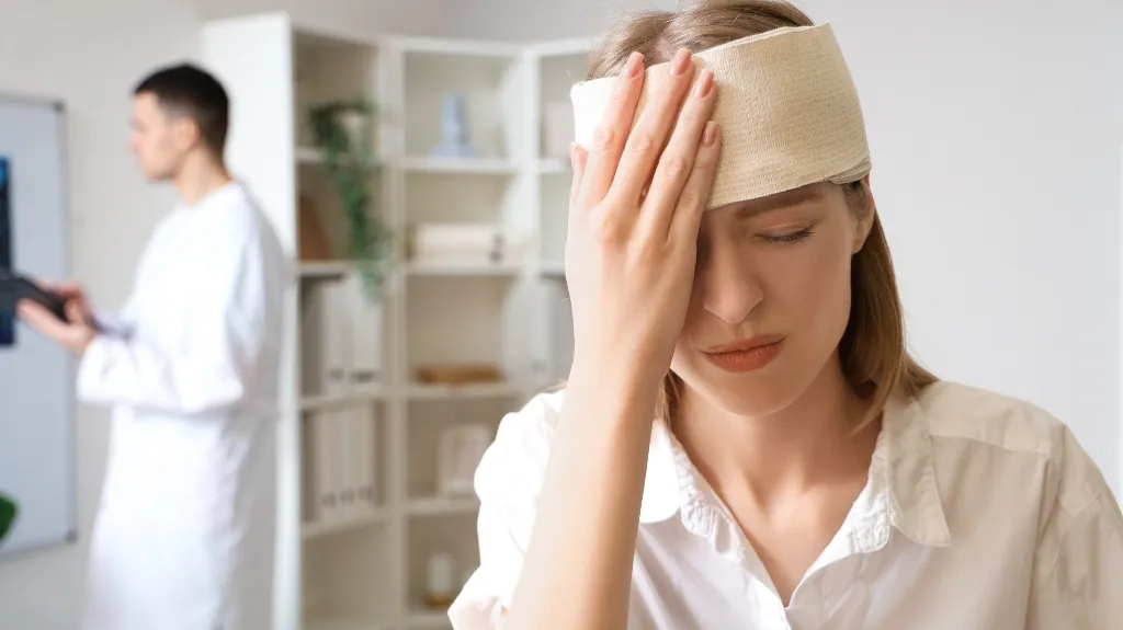 Woman with a forehead bandage holding her head, man in white coat blurred in background.