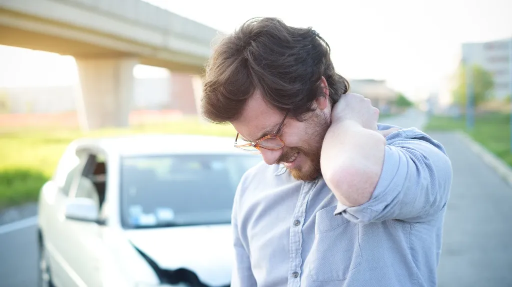 Man in glasses and light blue shirt holding neck in pain beside damaged white car on road.